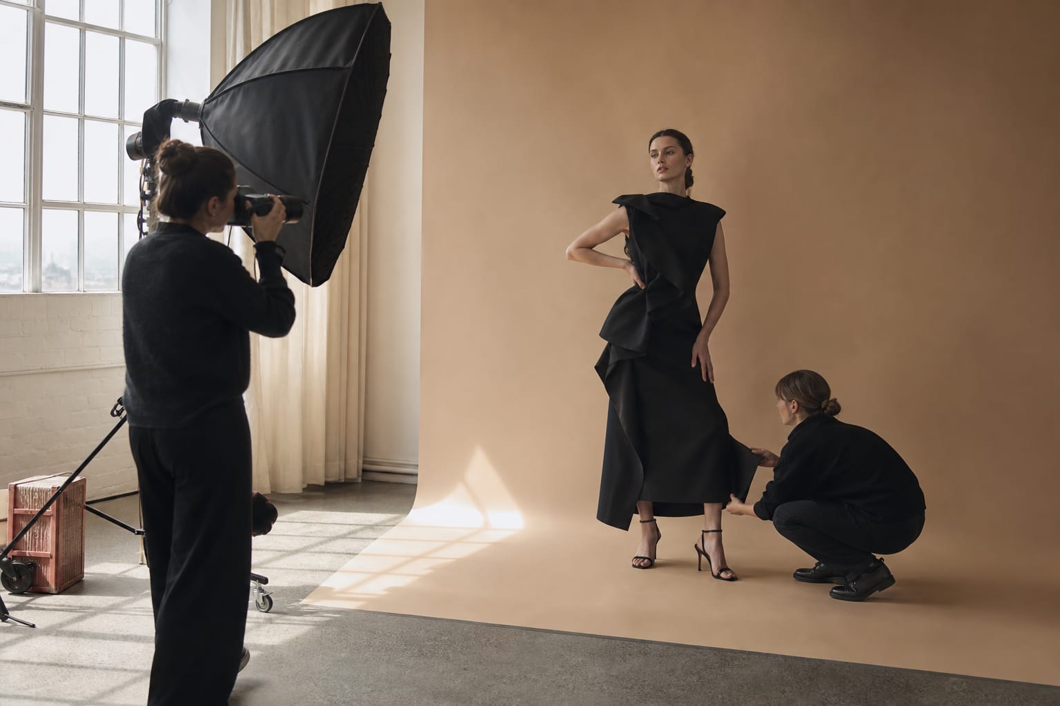 Žurnalo mados fotosesija studijoje: modelė juoda skulptūriška suknele pozuoja, šalia fotografas ir stilistė