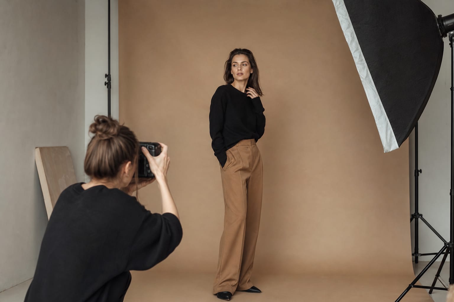 Fotomodelis studijoje pozuoja fotografui, šalia šviesos stovas ir šiltas smėlio spalvos fonas