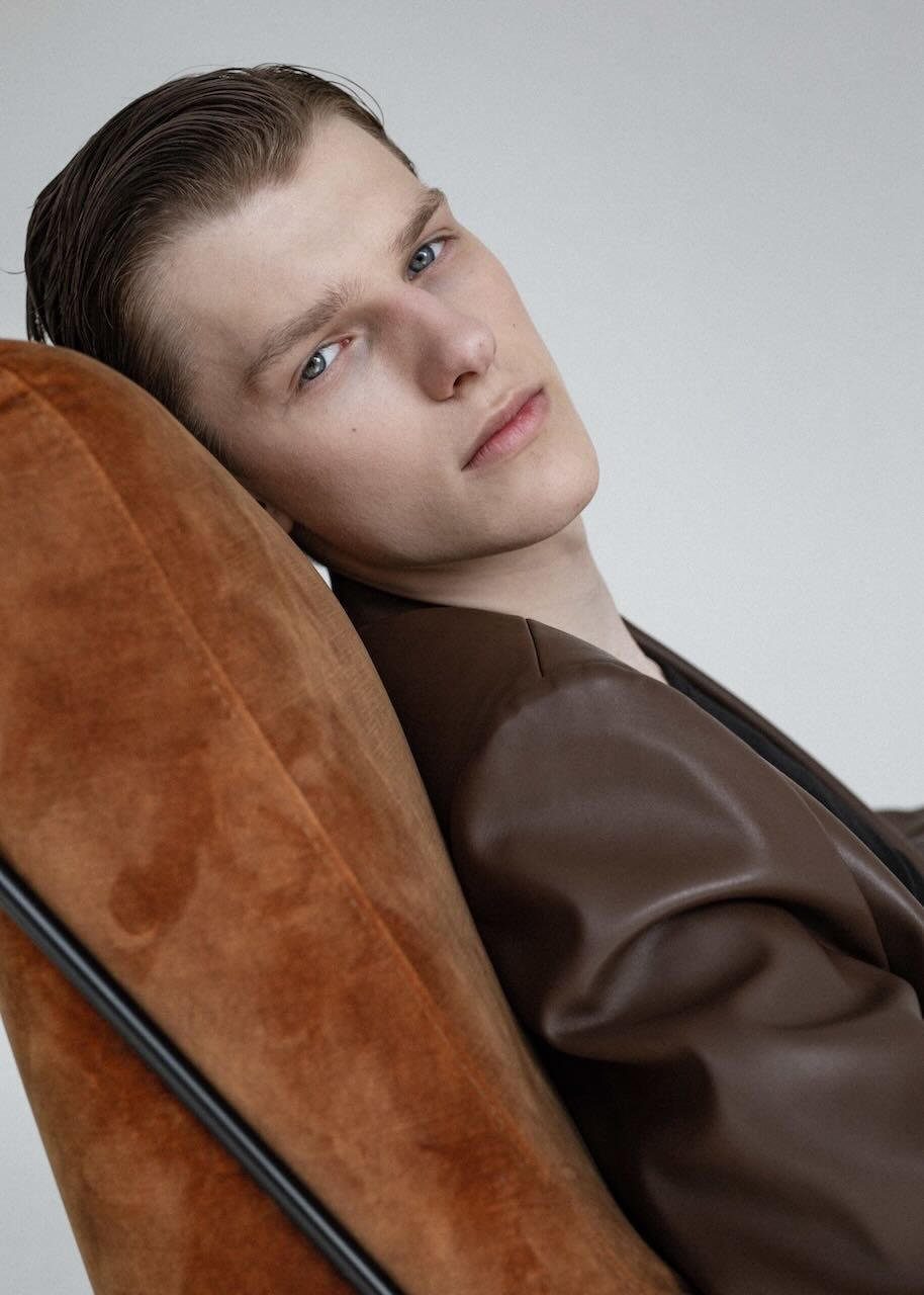Vyras modelis „Supermodels“ fotosesijoje Jaunas vyras modelis studijoje rudu odiniu švarku atsirėmęs į kėdę