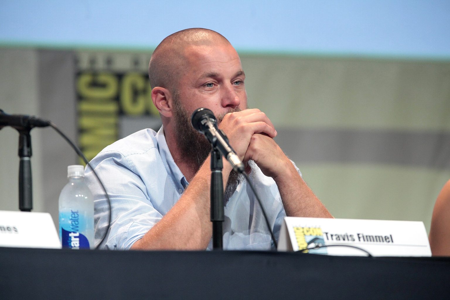 Travis Fimmel San Diego „Comic-Con“ 2015 m. Travisas Fimmelis nuskusta galva ir ilga barzda sėdi prie diskusijos stalo San Diego „Comic-Con“ 2015 m.