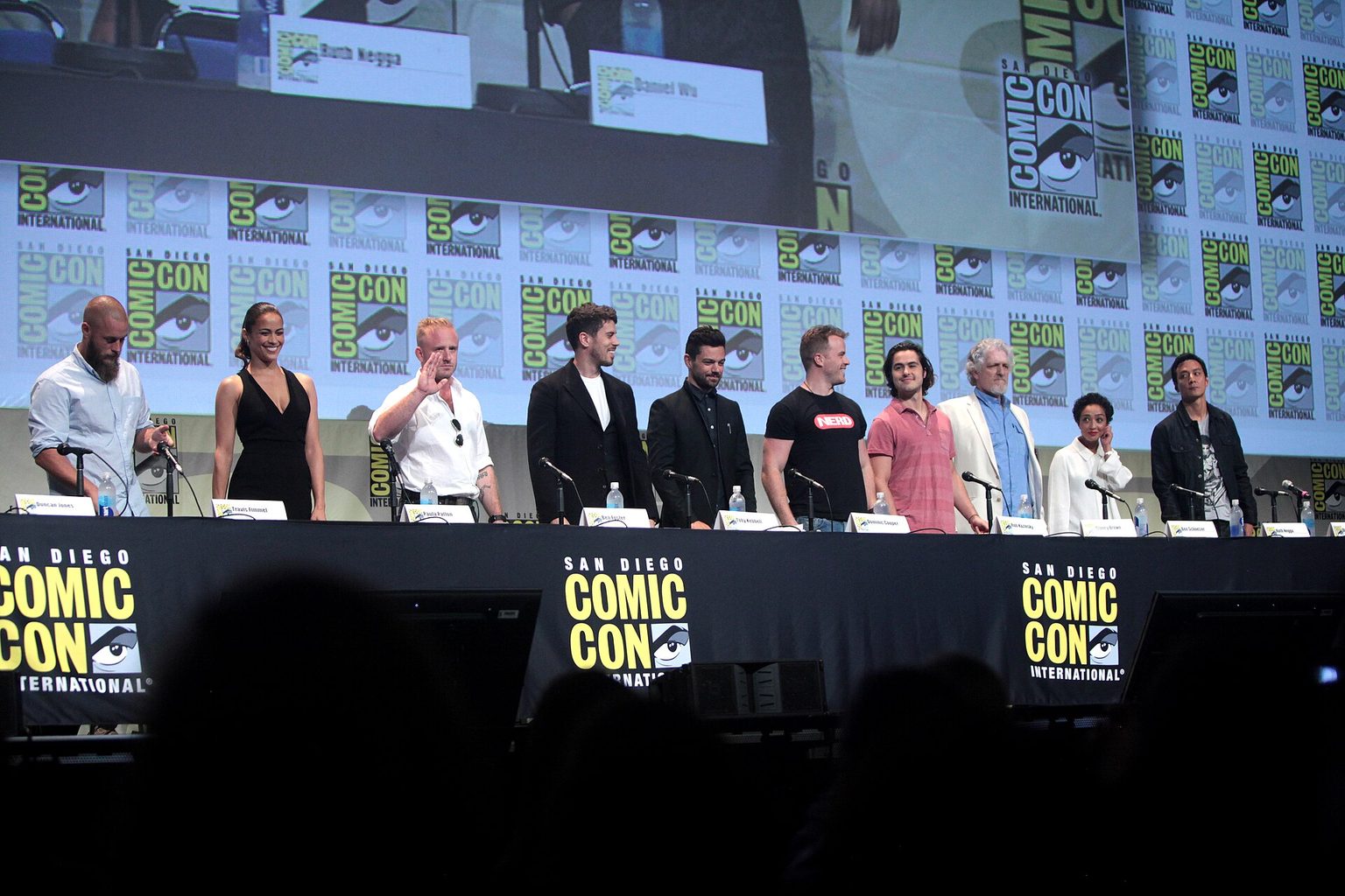 „Warcraft: Pradžia“ aktoriai San Diego „Comic-Con“ 2015 m. Filmo „Warcraft: Pradžia“ aktoriai stovi už ilgo stalo „Comic-Con“ scenoje 2015 m.